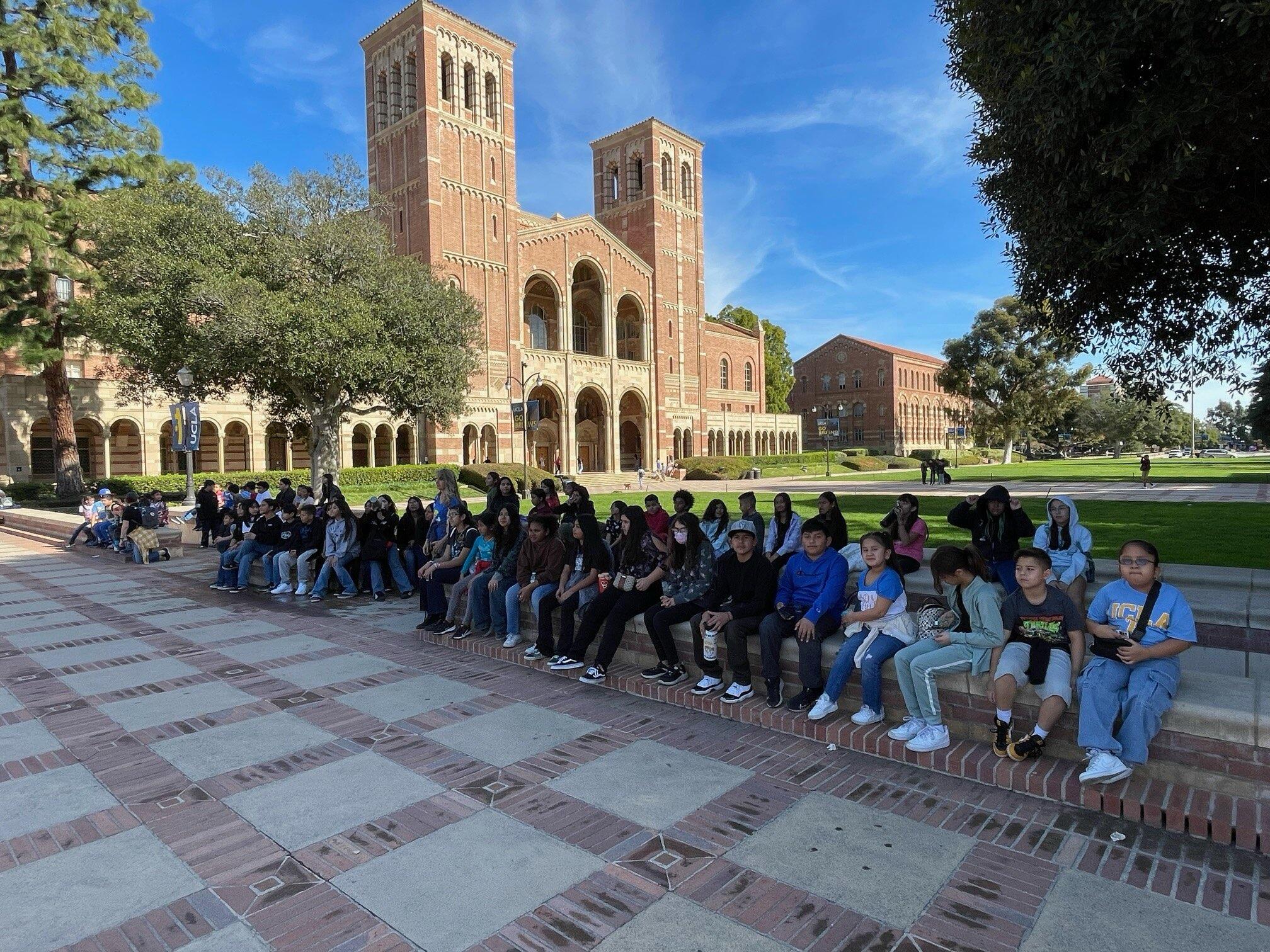 A diverse group of students are seated on steps outdoors, with a large, impressive brick building featuring arched doorways and a tower in the background, reminiscent of collegiate Gothic architecture. The students, mostly wearing casual clothing, seem to be on a campus tour or a school trip, with some looking at the camera and others enjoying the sunny day. The expansive lawn and clear skies suggest a peaceful, academic setting.