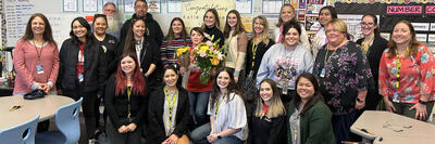 A joyful group of people gather for a group photo in a colorful classroom. The individuals, a diverse mix of ages and styles, are smiling broadly. Some are standing and others are seated at desks. They're casually dressed, most in work attire, suggesting they are colleagues. In the center, a woman holding a bouquet of flowers is presumably the focus of the celebration. The walls are covered with educational charts and student work, creating a lively and educational atmosphere.