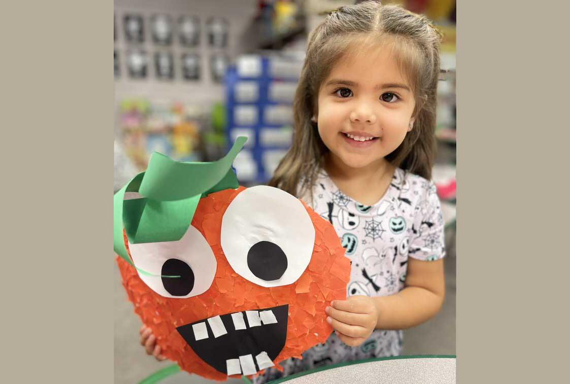 A young girl with a bright smile is holding a handmade paper lantern decorated to look like a jack-o'-lantern, with a green stem on top. She is wearing a white shirt adorned with Halloween-themed graphics. The background is blurred, with a classroom setting that includes storage bins and other educational materials.