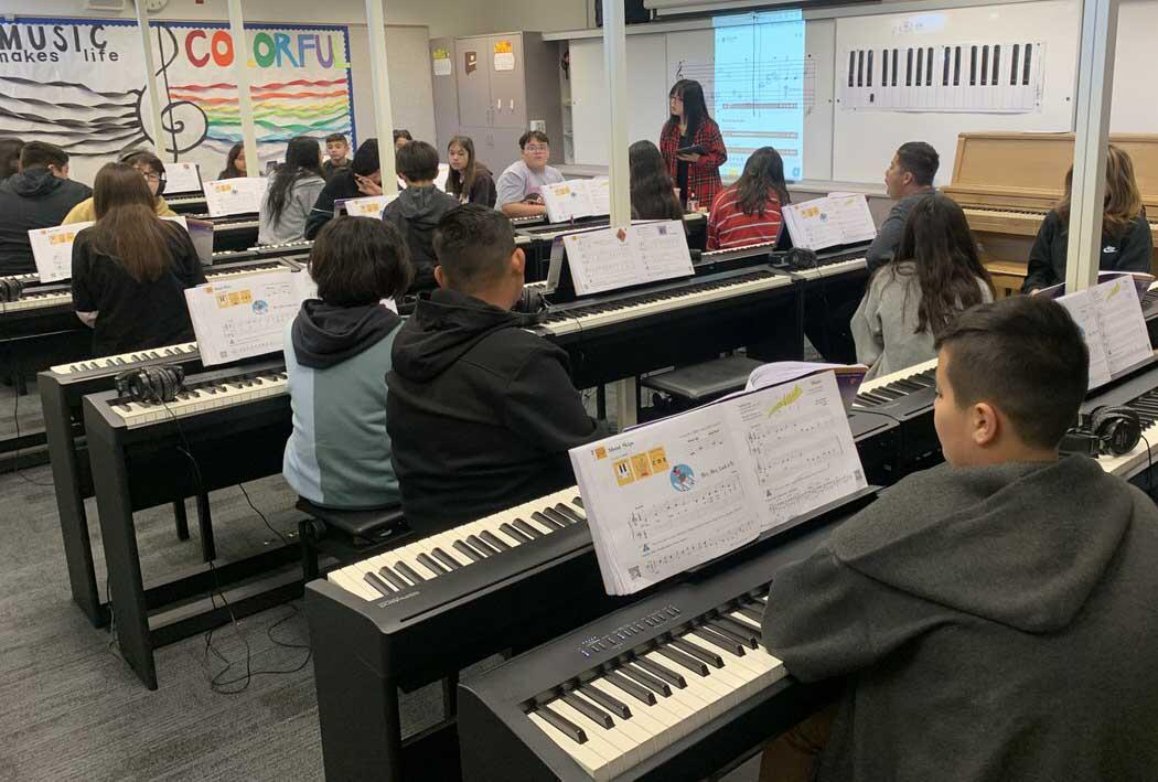 A classroom filled with students seated at digital pianos, focusing on their sheet music and the teacher's instructions. The teacher stands at the front, engaging with the students. The room has educational posters on the walls, including one that says 'MUSIC makes life COLORFUL.' This setting illustrates a music class in session, with students practicing their keyboard skills.
