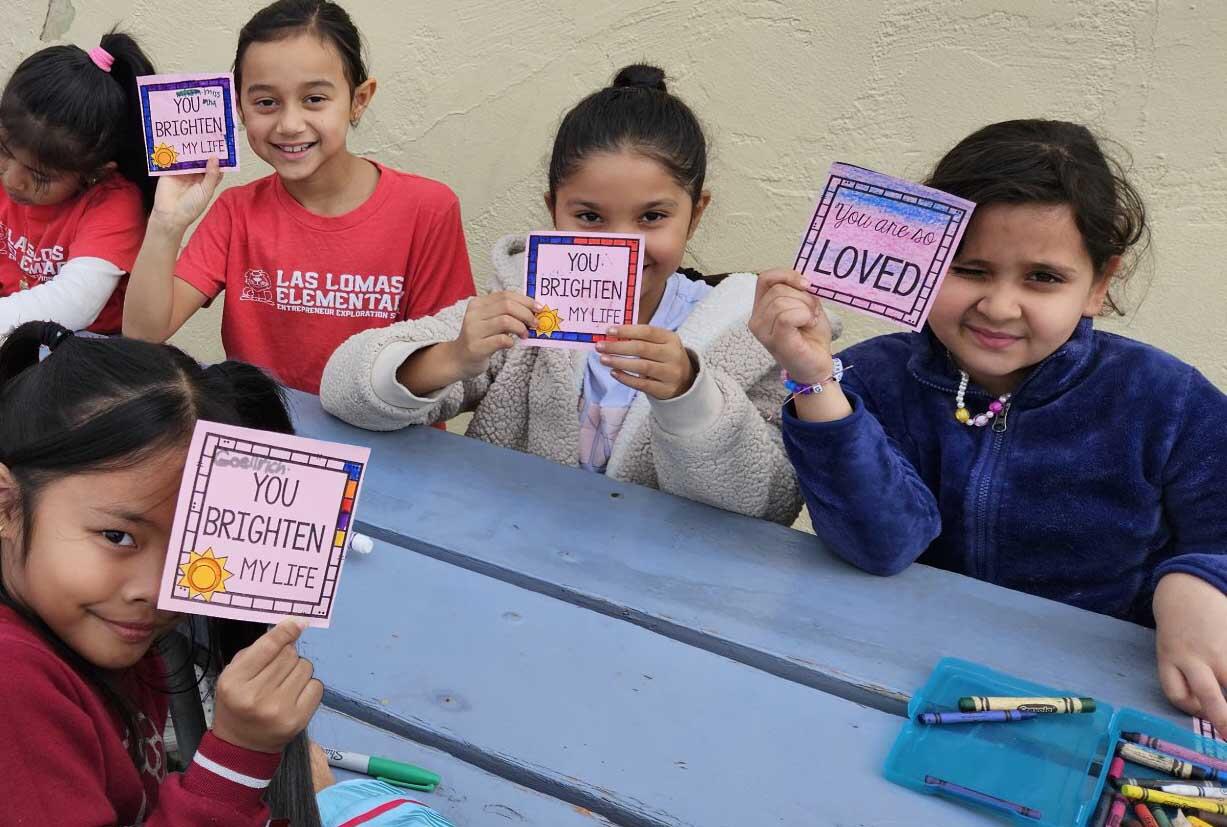 A group of smiling children sitting at an outdoor table, proudly holding up colorful handcrafted cards with positive messages such as 'You Brighten My Life' and 'You are so Loved.' One girl in the foreground is in a red 'Las Lomas Elementary' shirt, while others are in casual attire. Their bright expressions and the cards they're displaying convey a cheerful and supportive atmosphere, likely part of a school activity focused on kindness and affirmation.