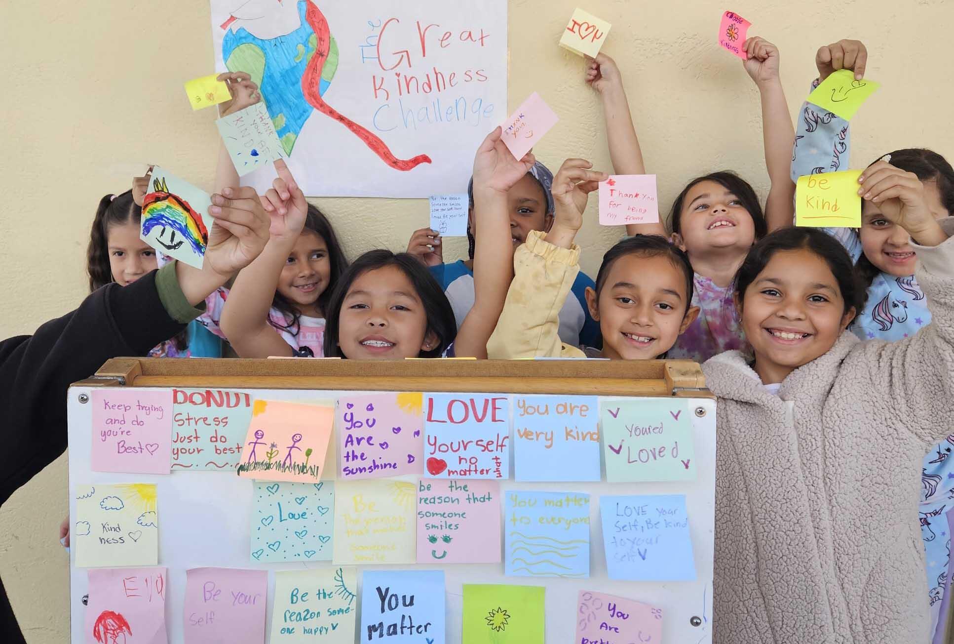 A group of exuberant children holding up colorful handmade notes and drawings in front of a poster that reads 'Great Kindness Challenge.' The children are showing off their messages of kindness and encouragement, with broad smiles on their faces. They stand in front of a bulletin board filled with similar notes, creating a vibrant and positive display in what appears to be a school setting.